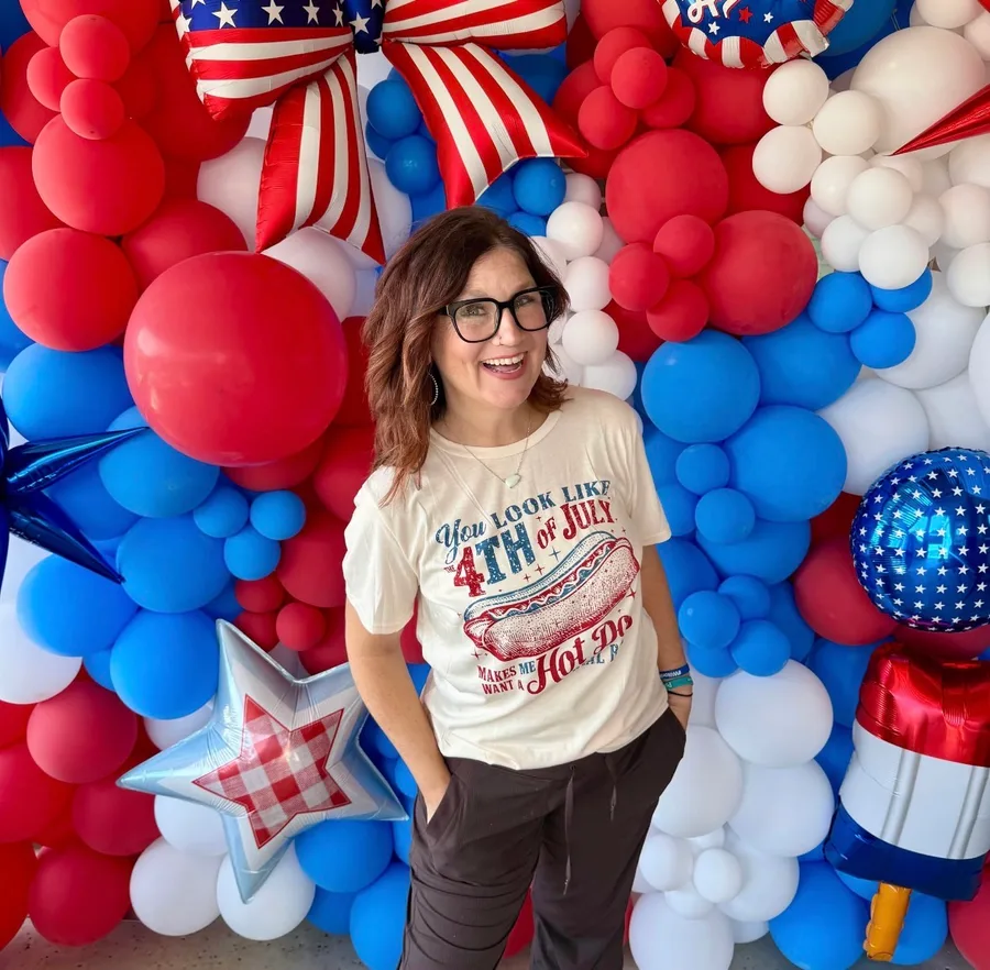 Becky, owner of Poppi Balloon Bar, smiling in front of one of her red, white, and blue balloon walls