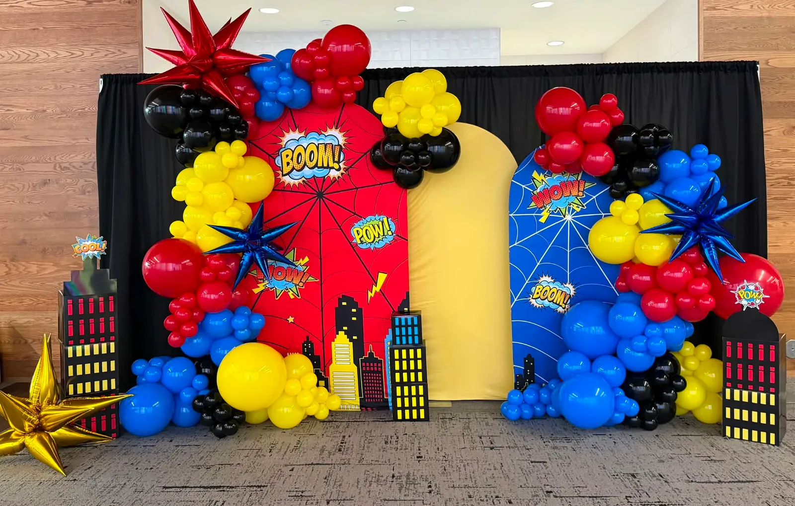 Comic-book superhero balloon setup with red and blue spiderweb arches, red, blue, yellow, and black balloons, foil starbursts, city-skyline cutouts, and BOOM, POW, and WOW signs