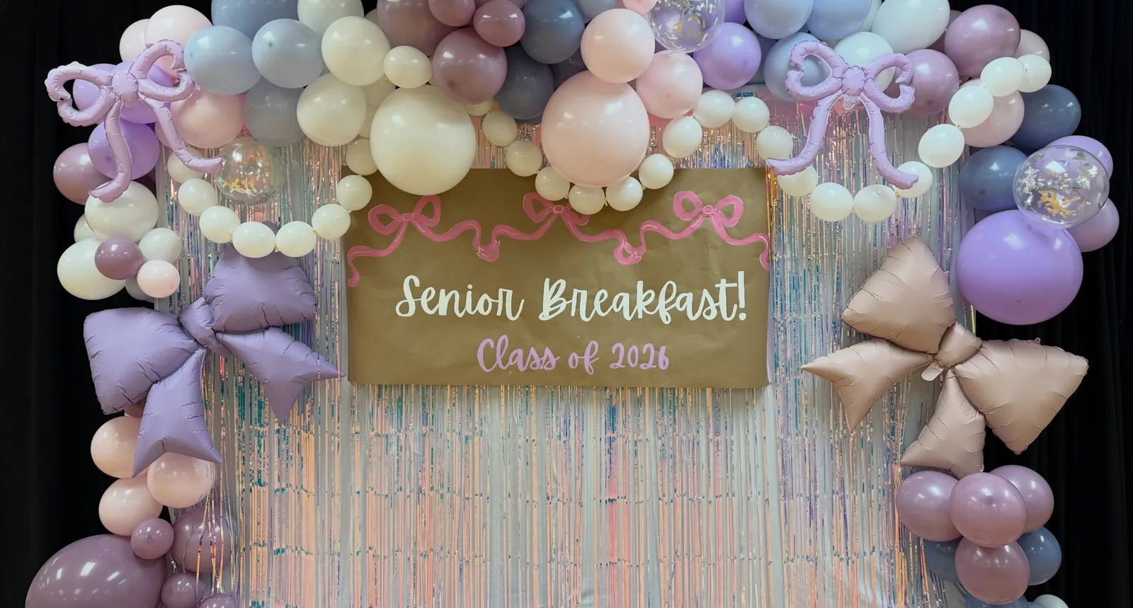 Pastel balloon wall in lavender, blush, and cream with foil bows framing a gold Senior Breakfast sign over a shimmer curtain